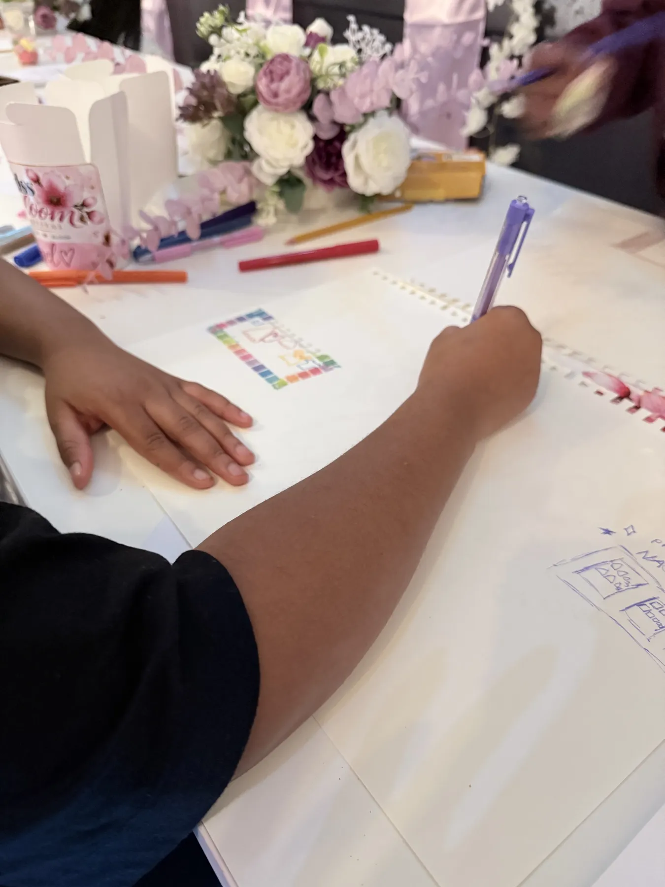 Handen van een meisje dat met een paarse pen een ontwerp tekent aan tafel.