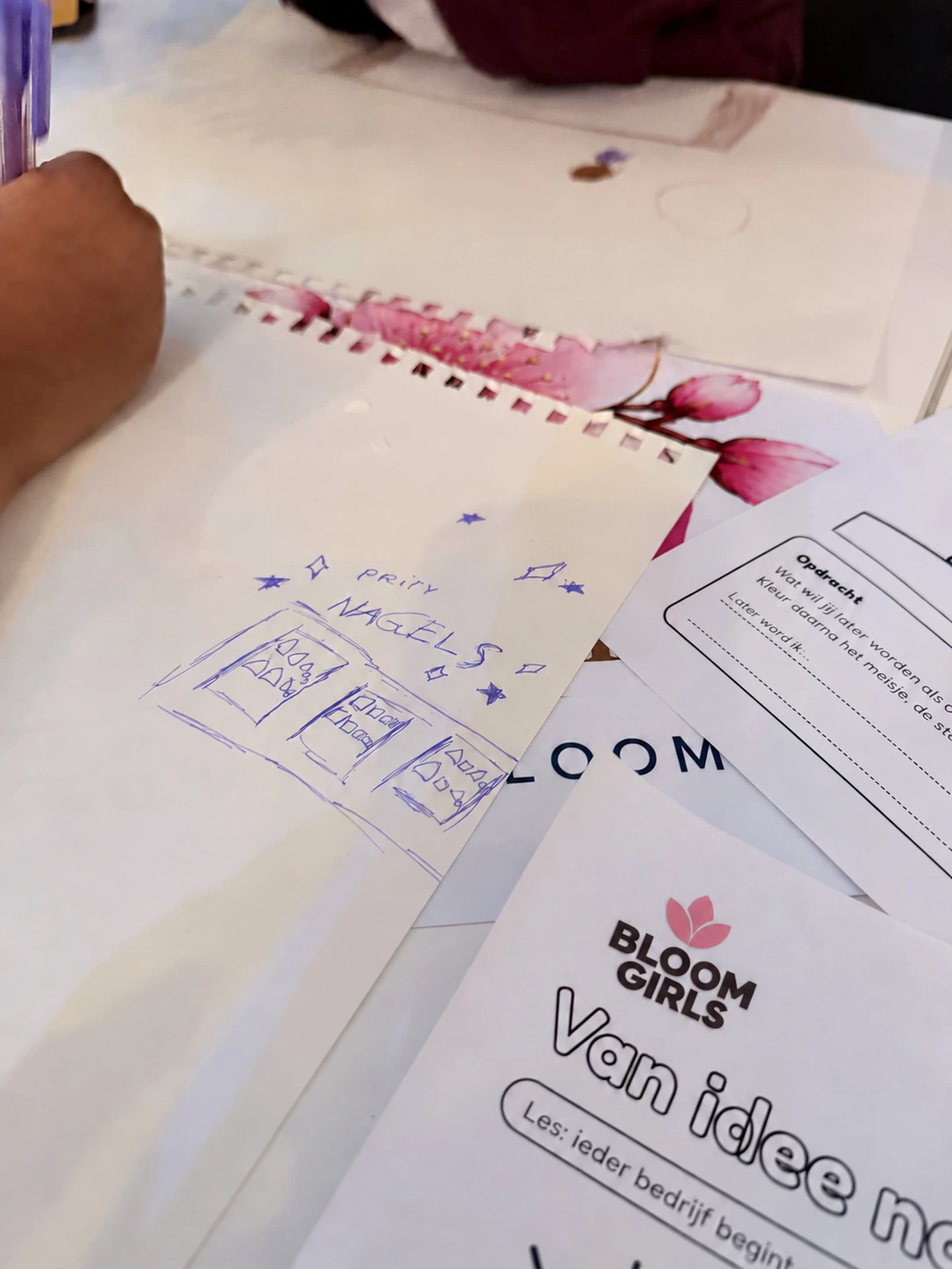 Een meisje tekent haar eigen idee voor een nagelstudio in een schetsboek, naast een Bloom Girls-werkblad over van idee naar eigen bedrijf.
