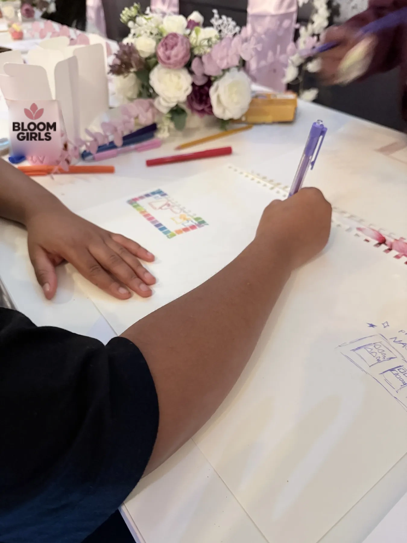 Handen van een meisje dat met een paarse pen een ontwerp tekent aan tafel.