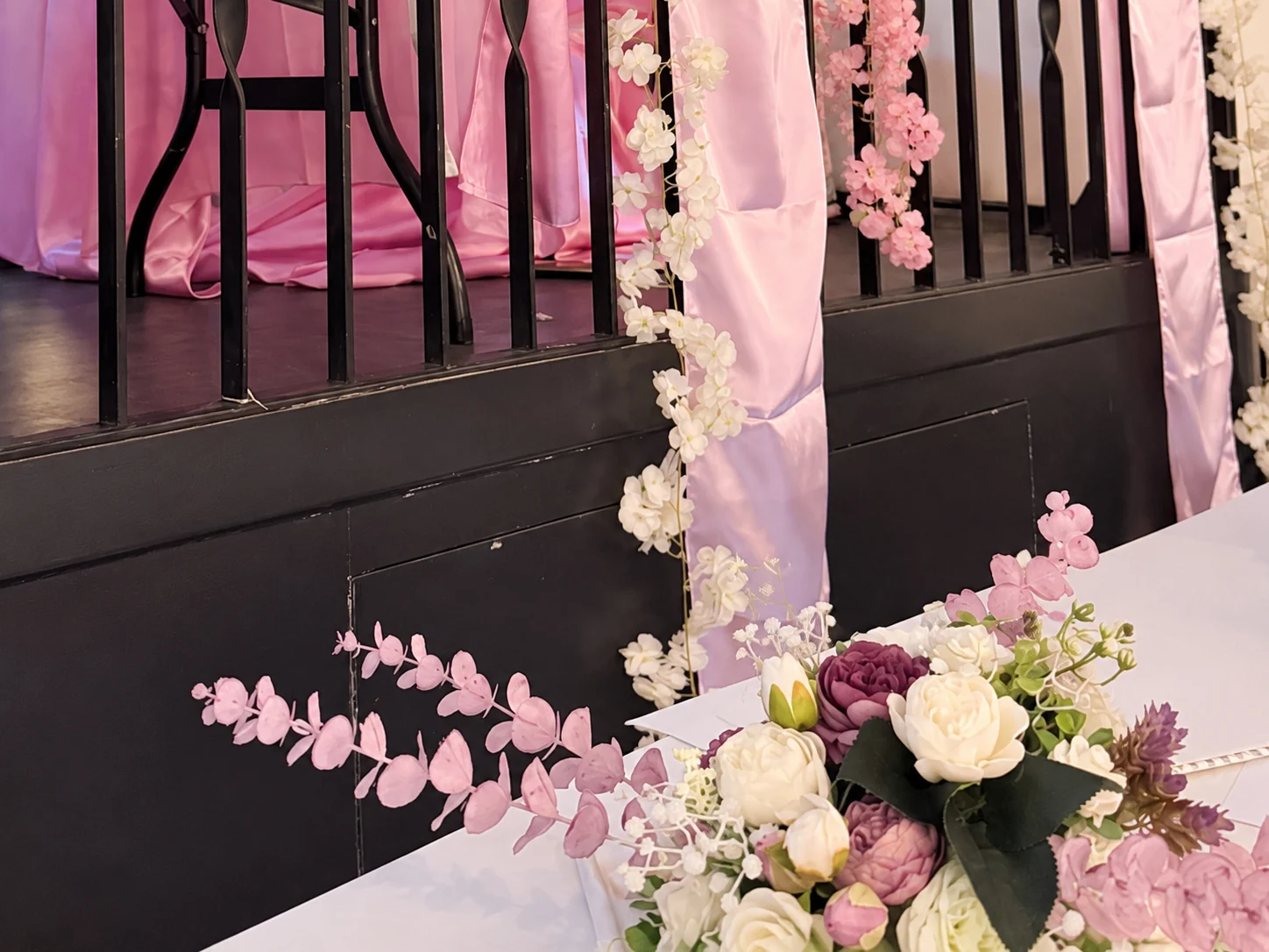 Detail van de aankleding: roze en witte bloemen, satijnen linten en een bloemstuk op de gedekte tafel bij het podium.