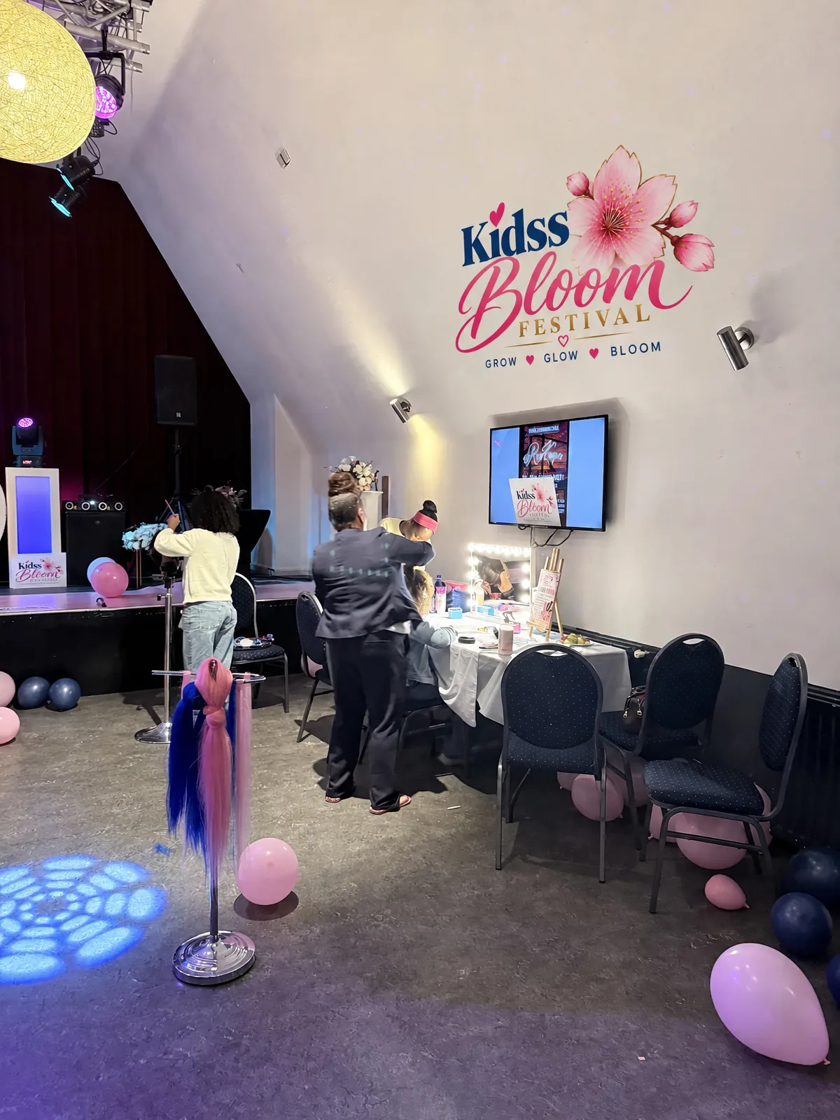 Een begeleider helpt een meisje aan de verlichte spiegeltafel van de beauty-hoek, met het Kidss Bloom Festival-logo op de muur.