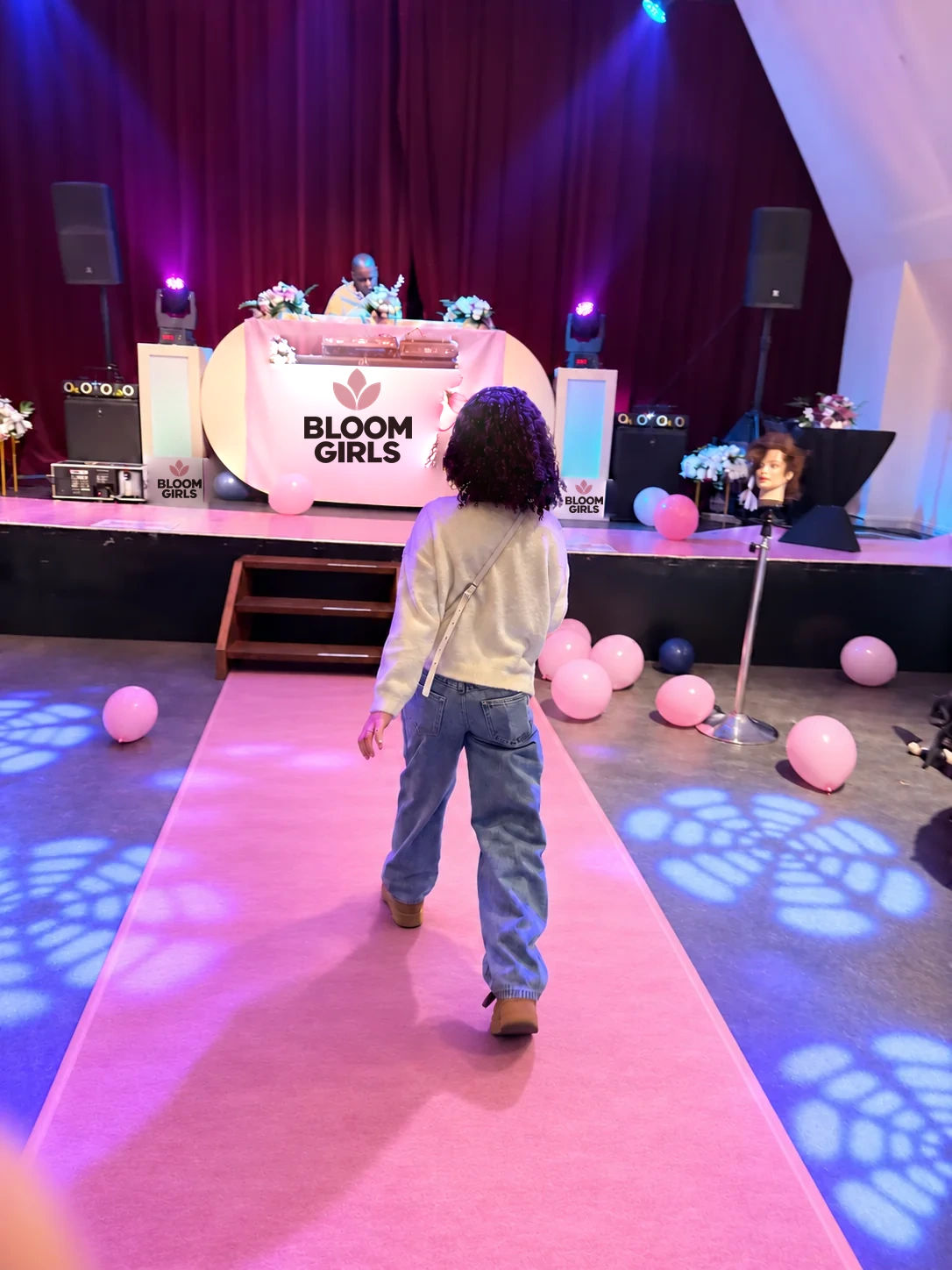 Een meisje loopt over een roze catwalk naar het podium, met paars en blauw licht op de vloer, de DJ op de achtergrond en het Bloom Girls-logo op het DJ-kleed.