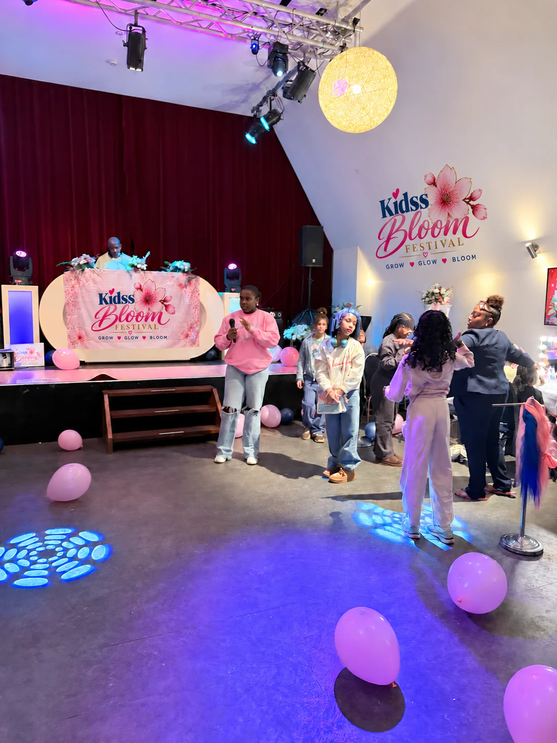 Een meisje spreekt met een microfoon voor de groep op de vloer voor het podium, met ballonnen en het Kidss Bloom Festival-logo op de muur.